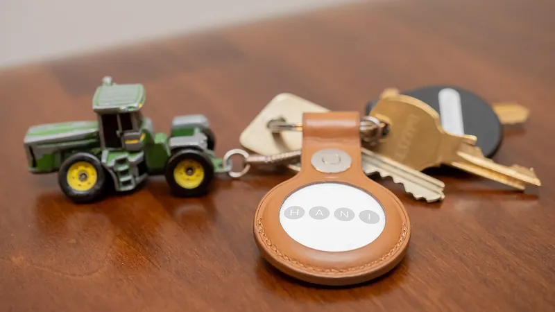 An Apple AirTag in a tan leather key ring holder lies on a wooden table next to keys and a small green toy tractor key chain