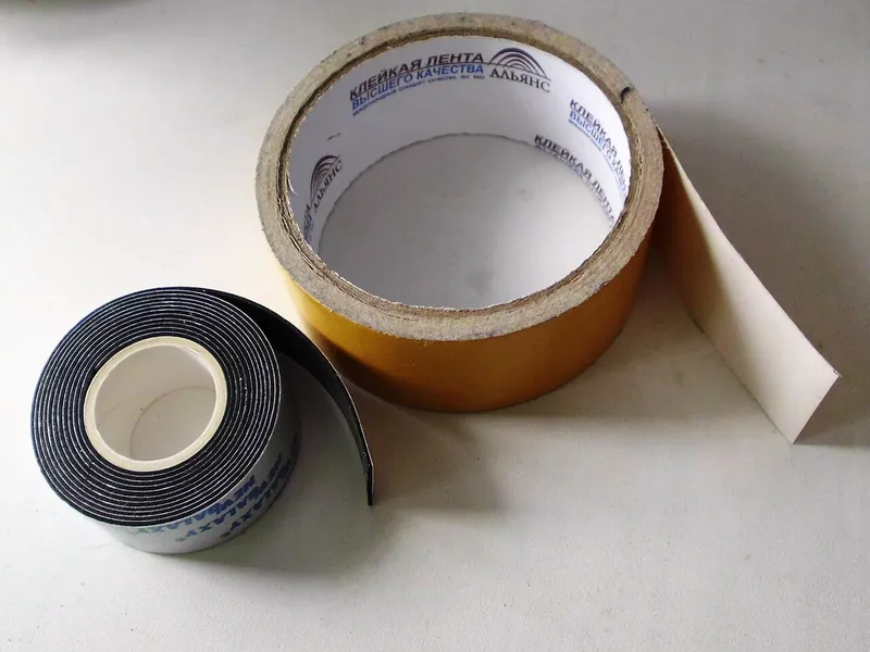 Two rolls of double-sided adhesive tape, one wide with a yellow liner and one narrow black foam roll, lying on a white table