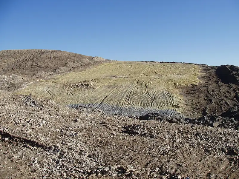 A bare hillside of pale yellow bentonite clay scarred by vehicle tracks under a clear blue sky, with grey rock in the foreground