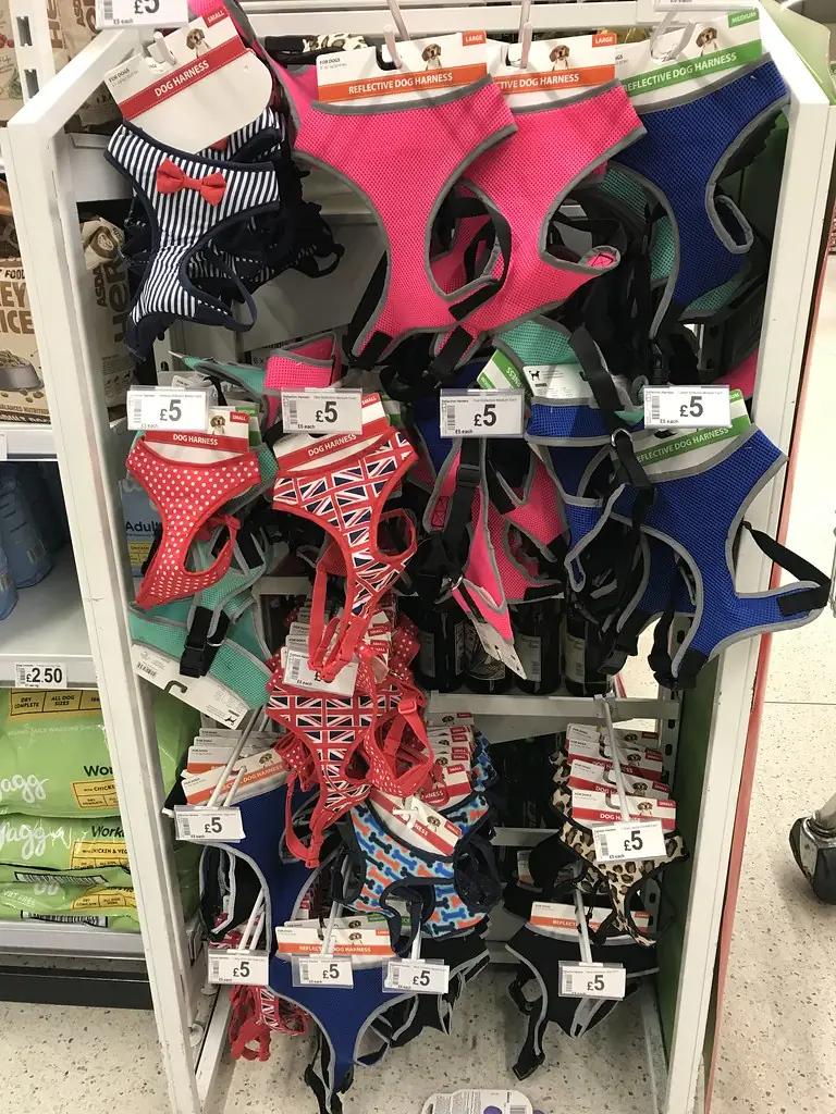 A pet shop display stand crowded with small padded dog harnesses in pink, blue, red and patterned fabrics, each with a price tag