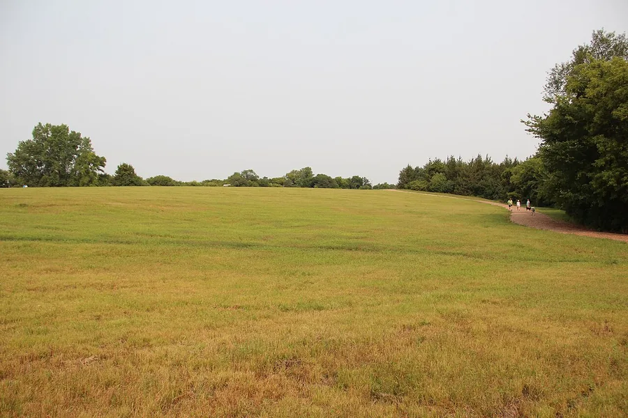 Wide grassy dog park under an open sky with trees at the edge and people walking a dog on a gravel path