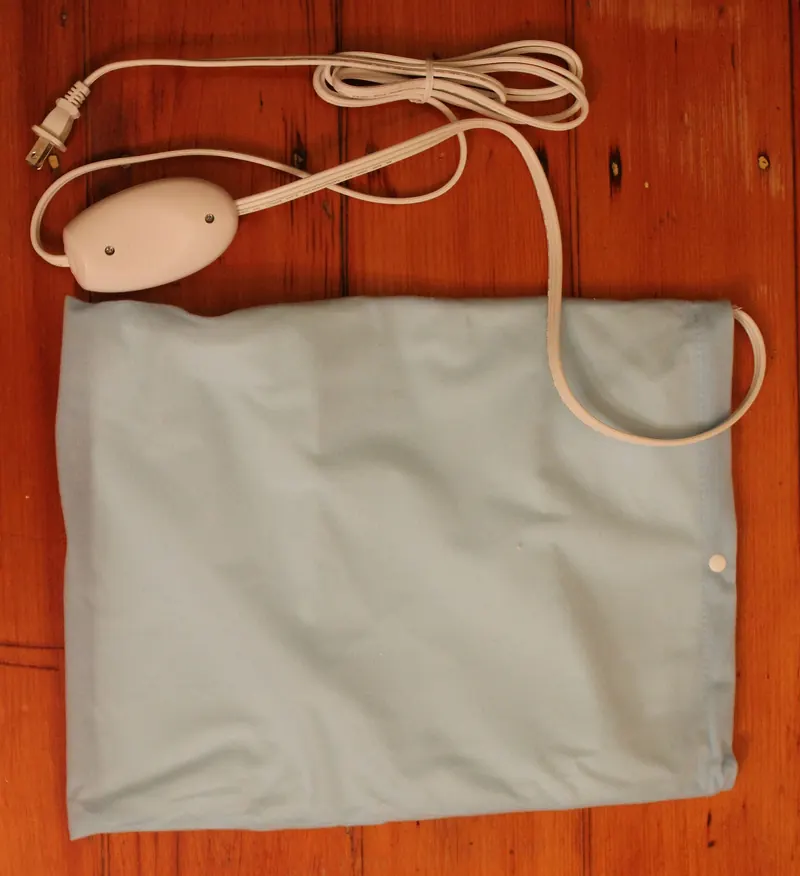 A light blue electric heating pad for people lies on a wooden floor with its white cord and hand controller coiled above it