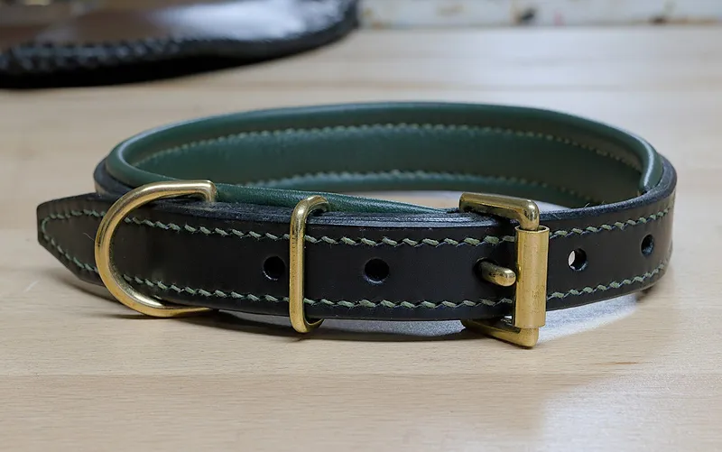A black leather dog collar with a green leather lining, green stitching, brass buckle, keeper and D ring on a wooden bench
