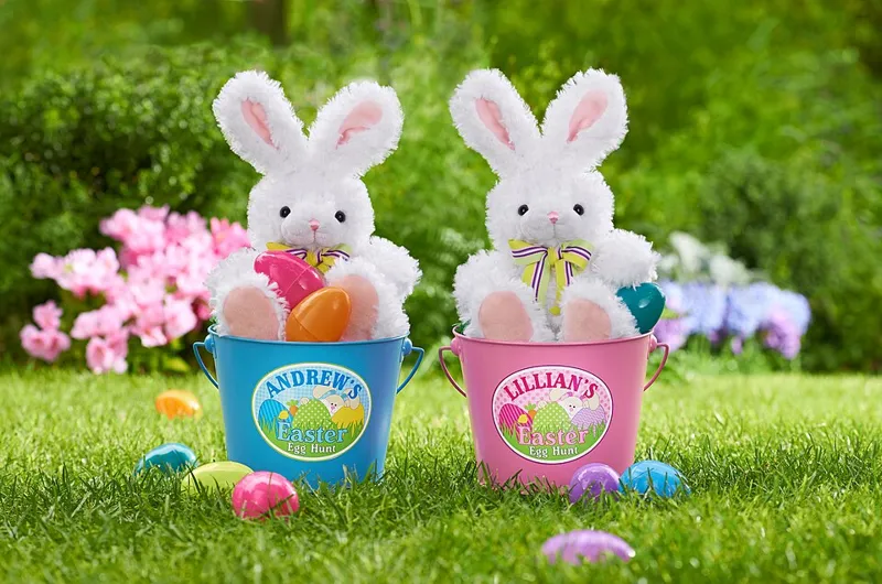 Two white plush bunnies in Easter buckets with plastic eggs on a lawn