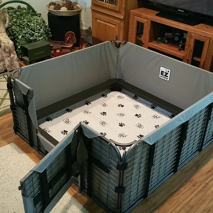 Empty modular whelping box with grey padded walls, a metal rail running around the inside, a fold-down door panel and a washable paw-print liner on the floor